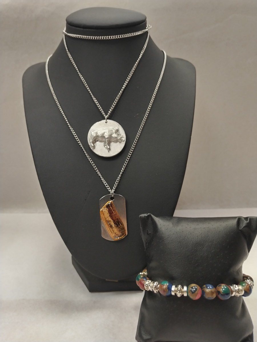 Trois pièces faites main sur un buste noir : un médaillon rond en étain où un sanglier apparaît gravé en relief, un pendentif rectangulaire de bois enrobé de résine sur plaque inox, tous deux montés sur chaîne argentée, et un bracelet de grosses perles irisées séparées par des rondelles strassées.